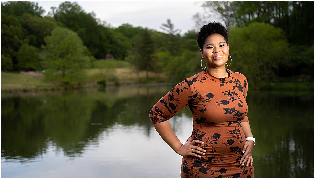 A black teen poses near a pond dressed in a brown dress with a black leaf pattern. 