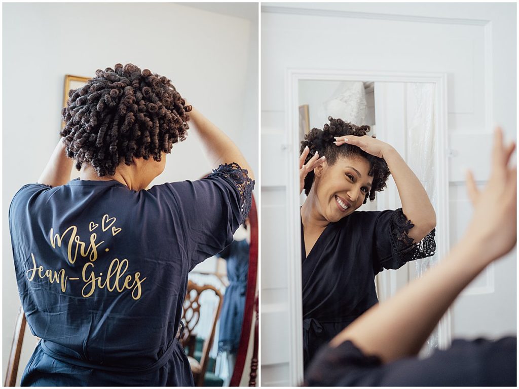 Bride wears a robe with her new last name on the back and smiles in the mirror while getting ready for her wedding at River Farm.