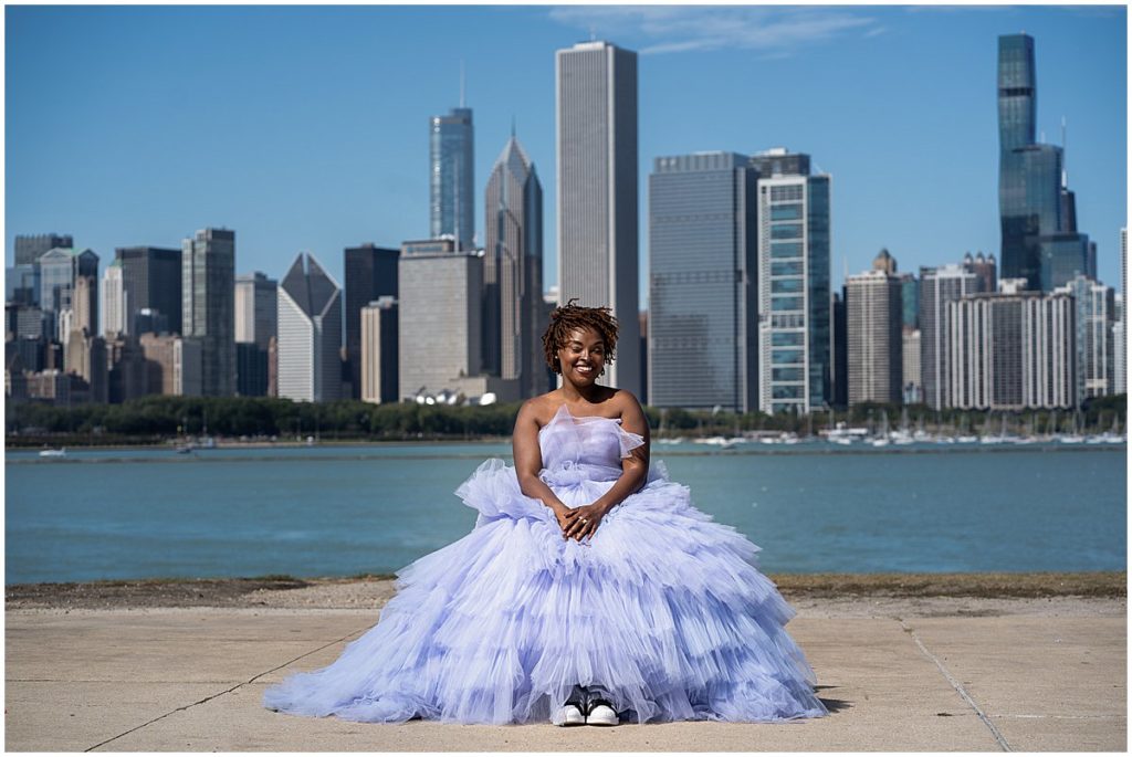 Black woman in a purple gown and sneakers sits on a rock with the Chicago skyline behind her.